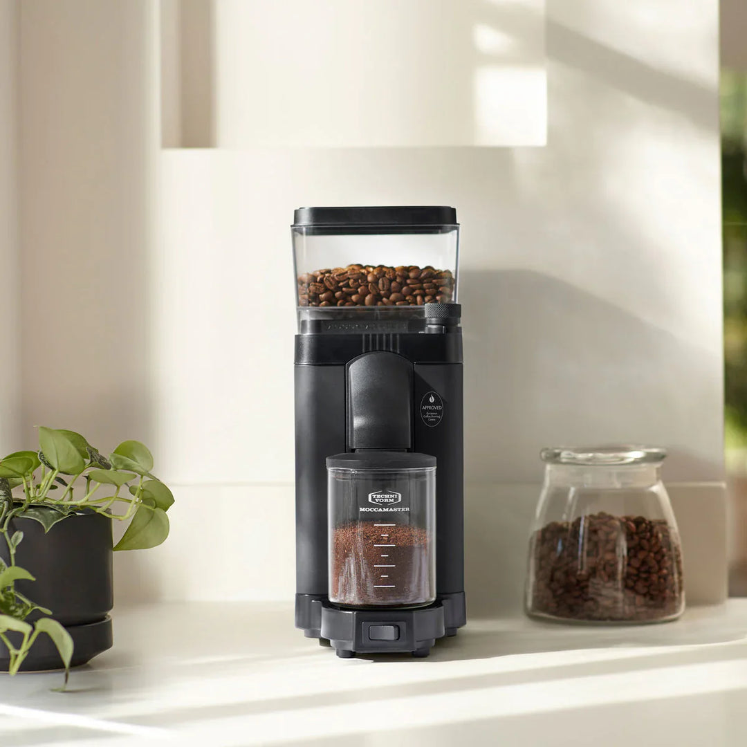 Moccamaster KM5 Coffee Grinder in Matte Black on a kitchen worktop next to a jar of coffee beans and a pot plant