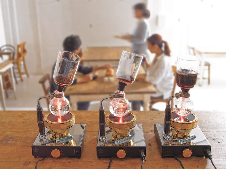 Three Hario Technica Coffee Syphon coffee makers on a wooden table with a blurred cafe background