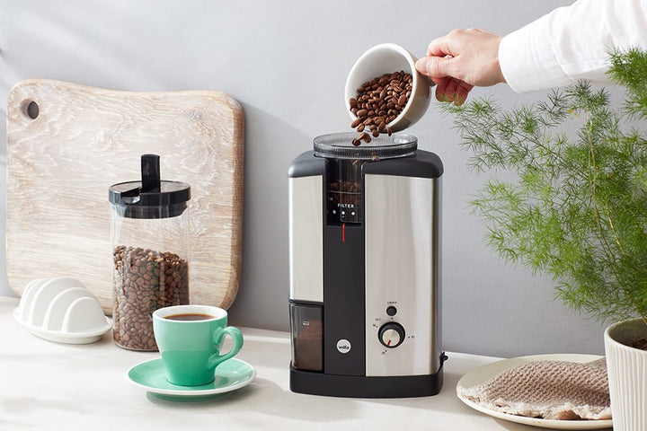 Person pouring coffee beans into a Silver Wilfa Svart Coffee Grinder on a kitchen counter.