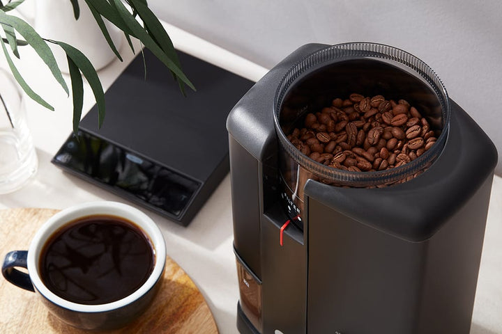 Wilfa Svart Aroma Precision Coffee Grinder in Black with coffee beans and a cup of coffee on a table.
