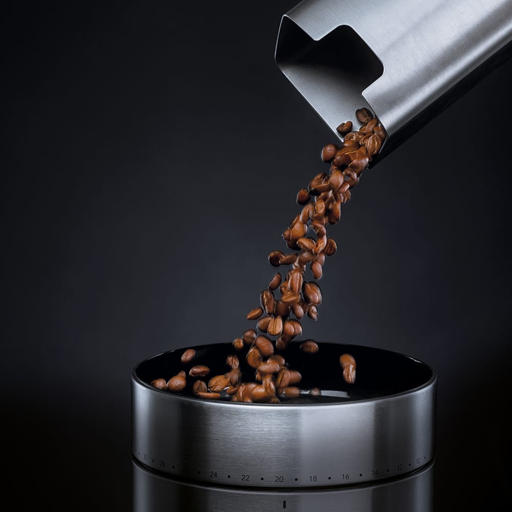Coffee beans being poured from a metal scoop into a Wilfa Uniform Stainless steel coffee grinder on a dark background