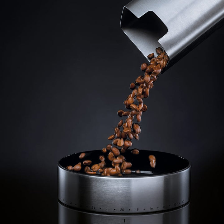 Coffee beans being poured from a metal scoop into a Wilfa Uniform Stainless steel coffee grinder on a dark background