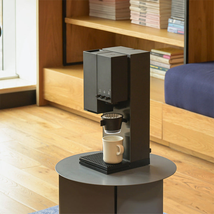 Midnight Black XBloom Studio Coffee Machine on a small round table with a cup underneath, in a room with wooden flooring and shelves.