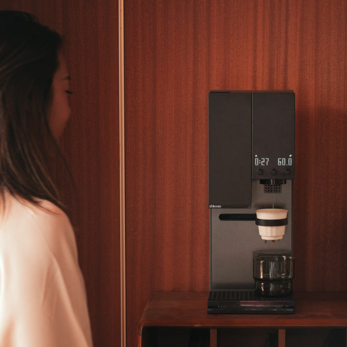 A woman standing next to a Midnight Black XBloom Studio Coffee Machine with a cup on a wooden wall.