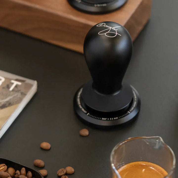 TIMEMORE FLOW self-leveling tamper held above a portafilter basket of ground coffee, espresso shot in the background