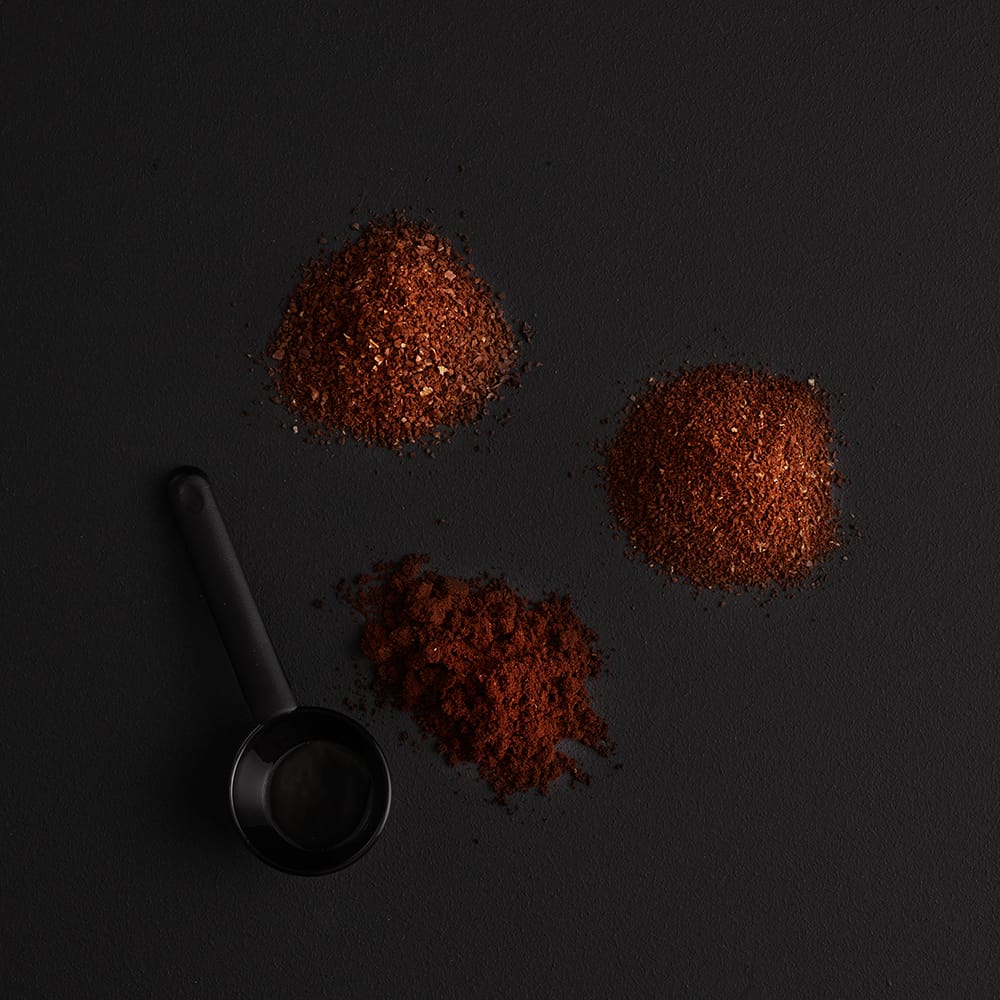 Three piles of ground coffee on a black background with a black scoop.