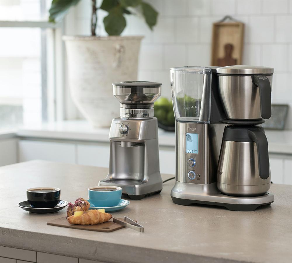 Coffee-making equipment, including a Silver Sage The Dose Control Pro Coffee Grinder and coffee maker on a kitchen counter with a cup of coffee and pastries.