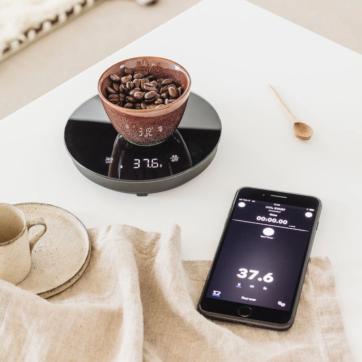 Black Wilfa Uniform Coffee and Kitchen Smart Scales with coffee beans and a smartphone app on a kitchen counter