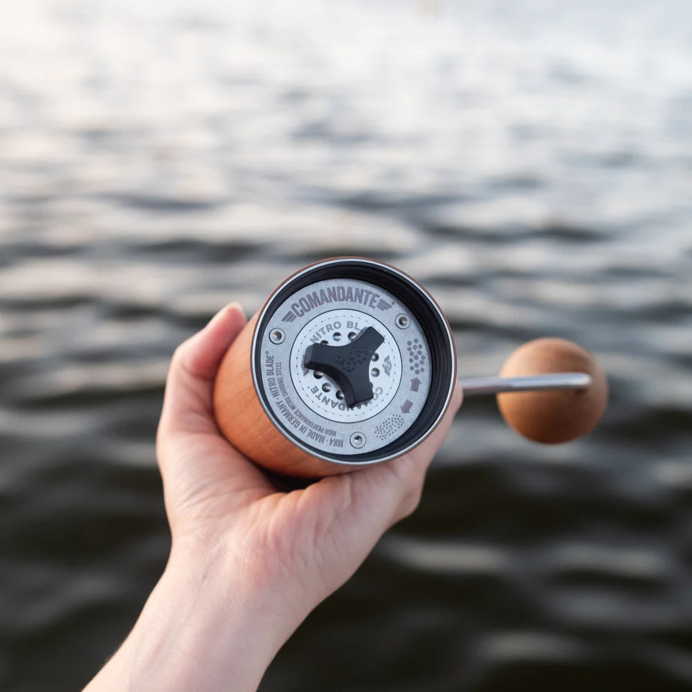 Hand holding a The Comandante C40 Nitro Blade Coffee Grinder MK4 in American Cherry with a blurred water background