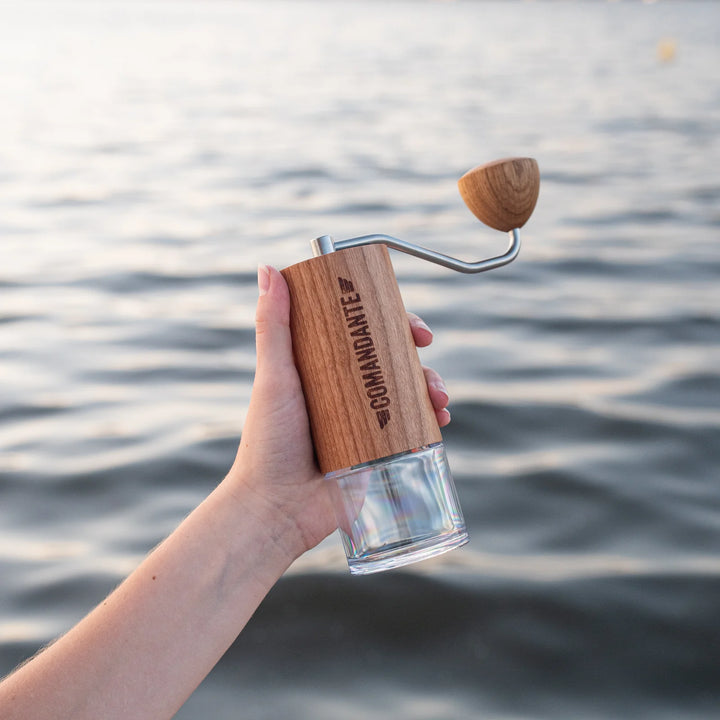 Hand holding a The Comandante C40 Nitro Blade Coffee Grinder MK4 in American Cherry with a clear glass base container against a water background