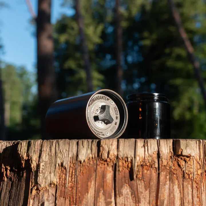Comandante C40 Nitro Blade Coffee Grinder MK4 in Liquid Amber with a Comandante brand logo on a wooden surface with trees in the background