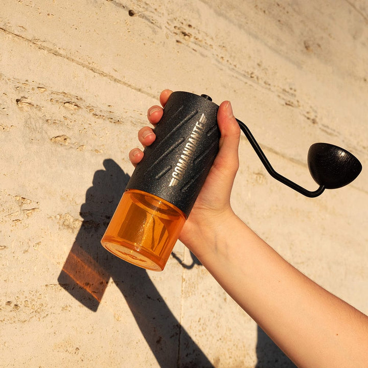 Hand holding a Comandante C60 Baracuda Coffee Grinder in Nautilus Blue against a beige wall.