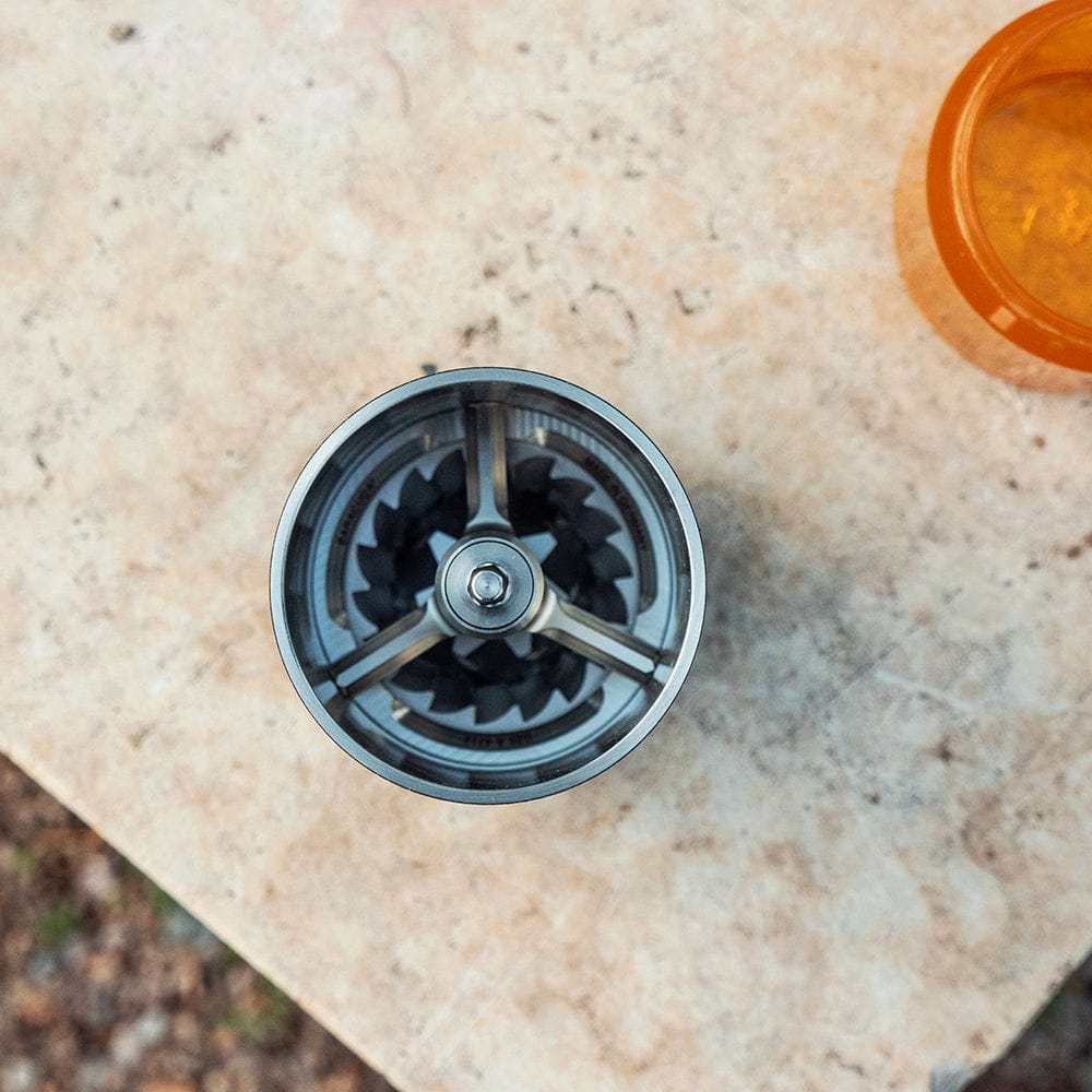 Top-down view of Comandante C60 Baracuda Coffee Grinder in Nautilus Blue on a beige stone surface with an orange coffee grounds container in the corner