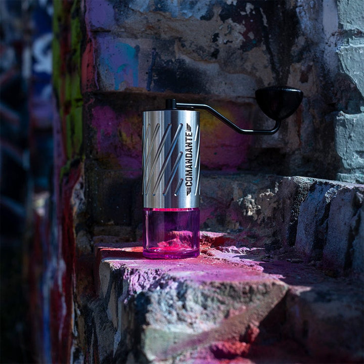 Comandante C60 Baracuda Coffee Grinder in Velvet Steel with pink bean jar on a textured surface