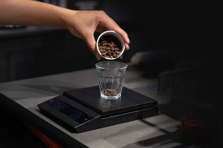 Hand pouring coffee beans into a glass on a Felicita Parallel Coffee Scale in Black with a dark background