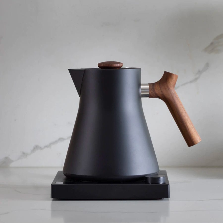 The Fellow Corvo EKG Electric Kettle in Matte Black with a Walnut wooden handle on a light marble background