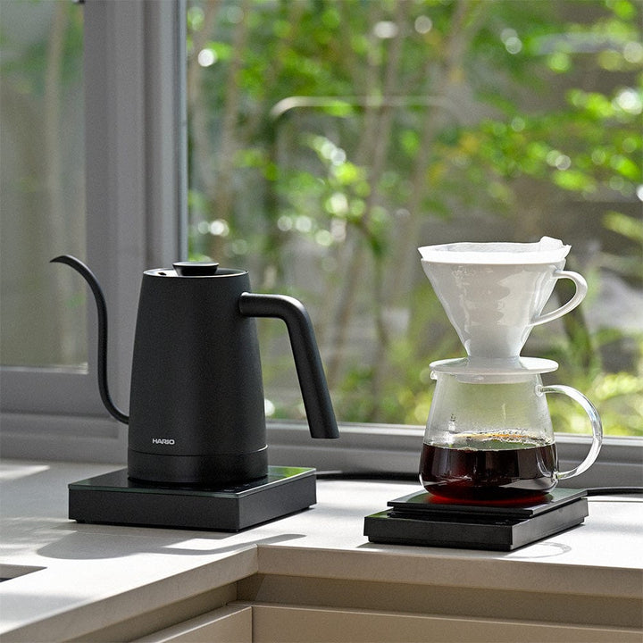 Black Hario Lyra electric kettle and glass coffee maker on a windowsill with a view of greenery.