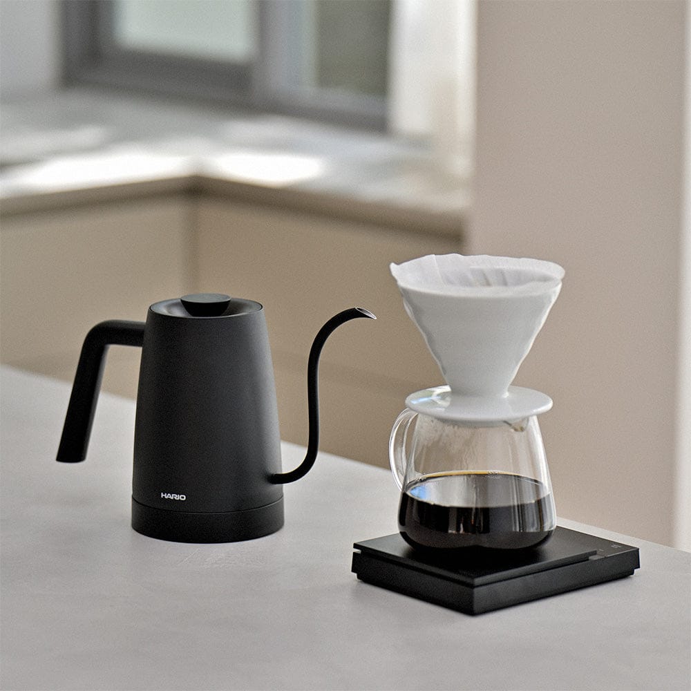Black Hario Lyra electric kettle and glass coffee maker with a white filter on a light surface.