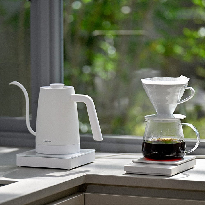 White Hario Lyra electric kettle and coffee maker on a windowsill with a blurred green outdoor background