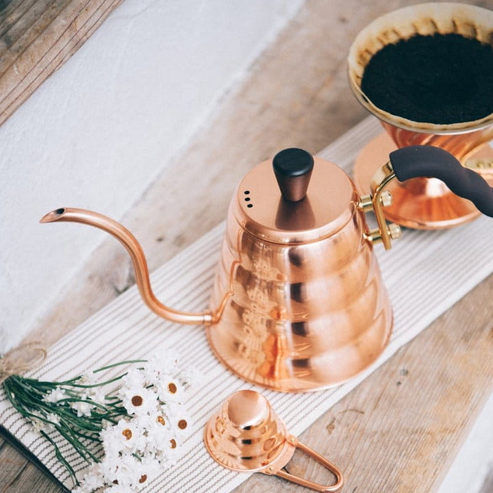 Hario V60 Buono Copper Stove Top Drip Kettle on a wooden surface with a white cloth and small plant.