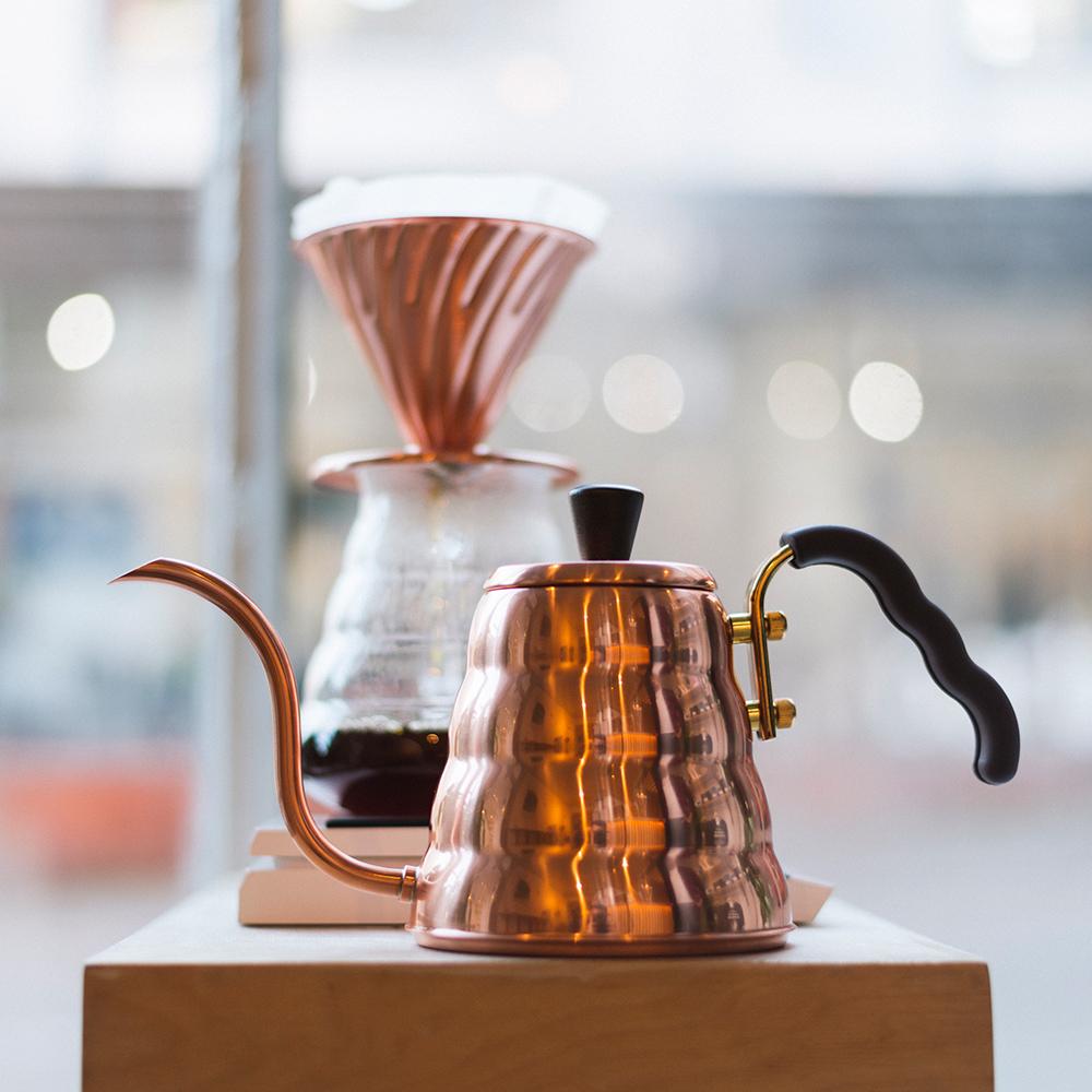 Hario V60 Buono Copper Stove Top Drip Kettle with a glass carafe on a wooden surface with a blurred background