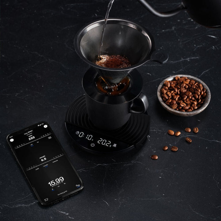 Coffee brewing process with a Black Wilfa Uniform Coffee and Kitchen Scale, phone app, and coffee beans on a dark surface.