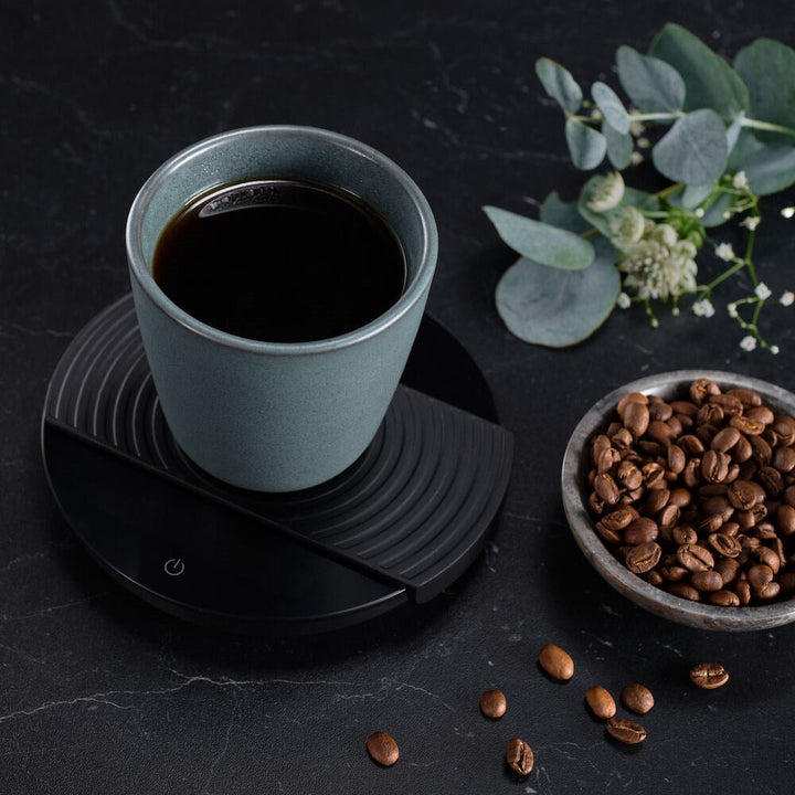 Teal mug with coffee on a Black Wilfa Uniform Coffee and Kitchen Scale, surrounded by coffee beans and greenery on a dark surface