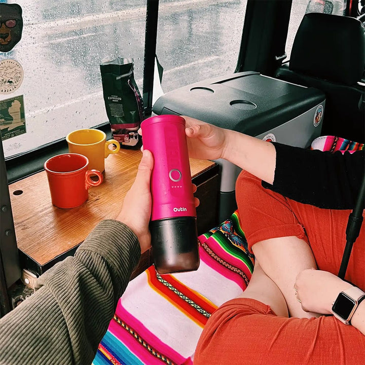 Person holding an OutIn Nano Portable Espresso Coffee Machine in Fuchsia Pink in a vehicle with coffee cups on a table.