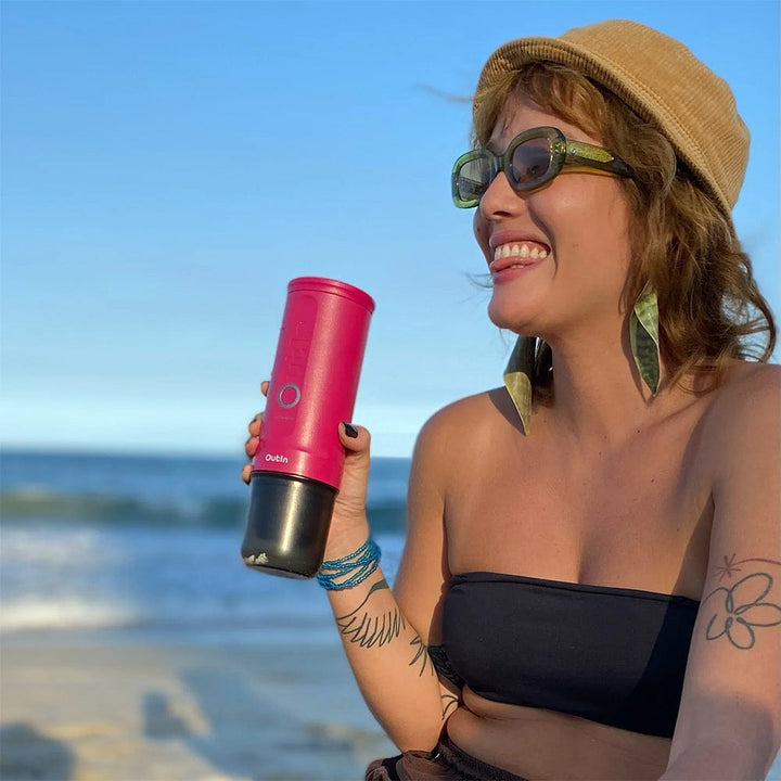 Woman on a beach holding a OutIn Nano Portable Espresso Coffee Machine in Fuchsia Pink with a blue sky and ocean background