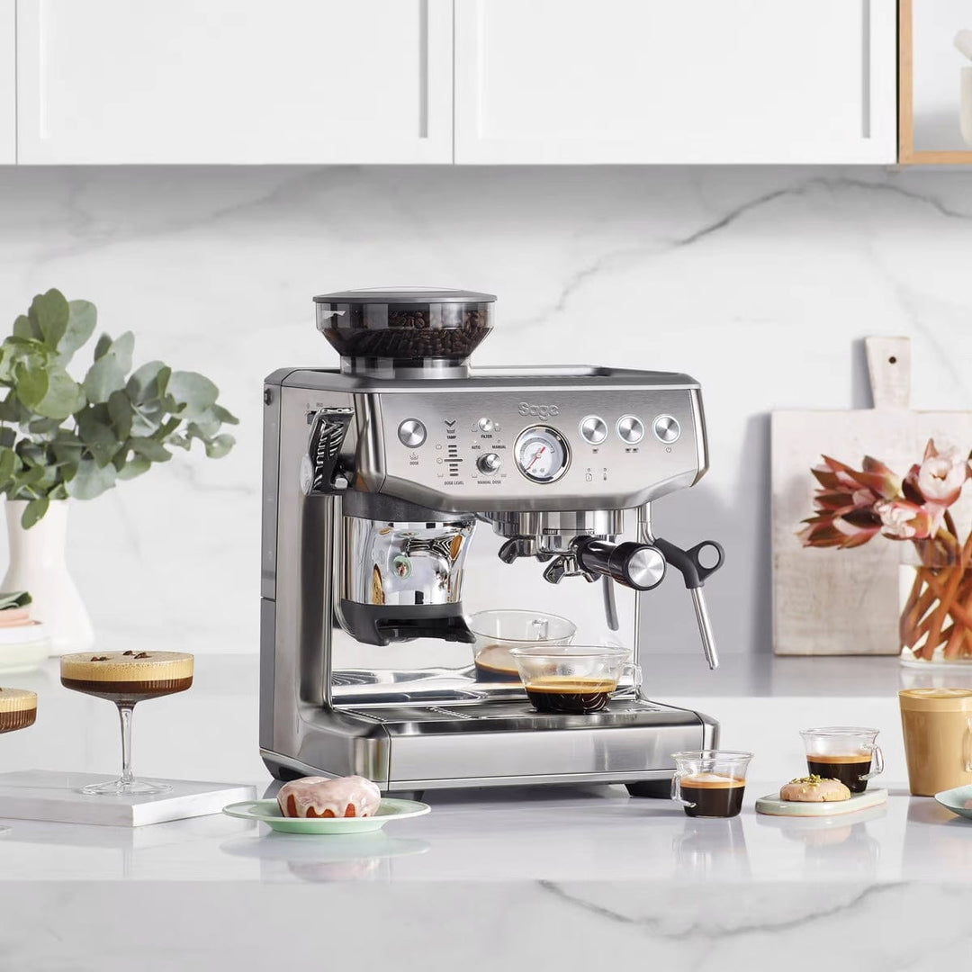Stainless steel Sage Barista Express Impress Espresso Machine on a kitchen counter with coffee and pastries.