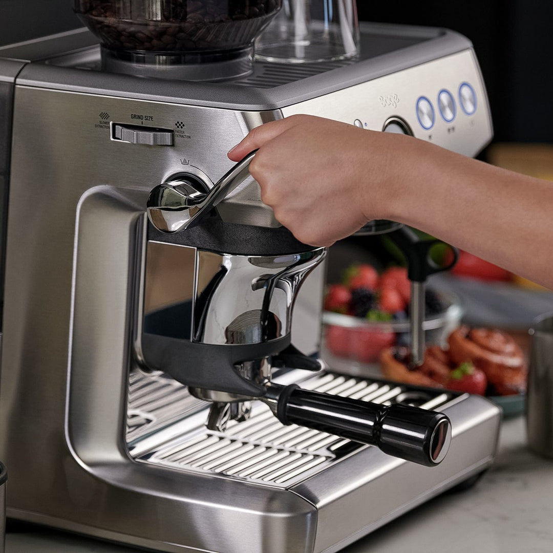 Person using a Stainless steel Sage Barista Express Impress Espresso Machine with a blurred background of fruits and kitchen items.