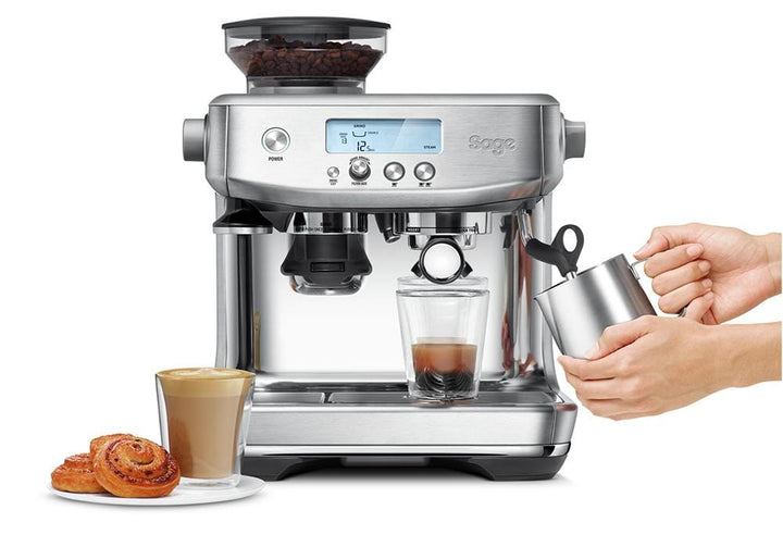 The Sage Barista Pro Espresso Machine in Stainless Steel with a glass of coffee and a hand holding a cup.