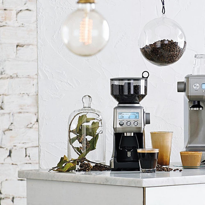 The Sage Smart Grinder Pro Coffee Grinder Black Truffle on a kitchen counter with coffee beans and a coffee machine in the background.