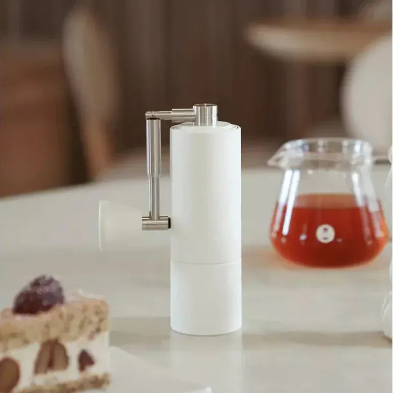 Timemore Chestnut C2 Manual Fold Coffee Grinder in White on a table with a carafe of fresh coffee and a piece of cake