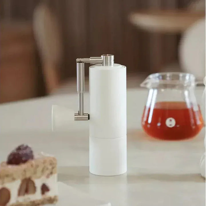 Timemore Chestnut C2 Manual Fold Coffee Grinder in White on a table with a carafe of fresh coffee and a piece of cake